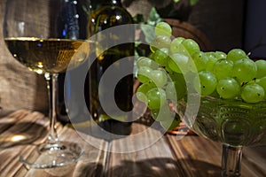 Two bottles of red and white wine on a wooden table with a bunch of grapes and a glass on a thin stem