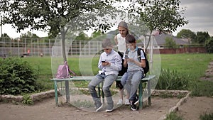 Two boys and girl use their phones during school breack. Cute boys sitting on the bench and play online games. Girl look