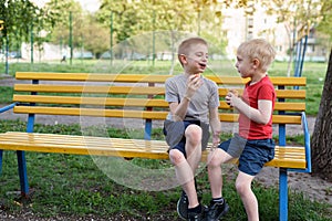 Two boys have a snack on a bench in the park and chat. Summer day
