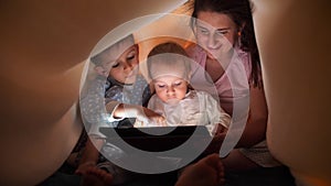 Two boys with mother hiding under bedsheet and playing games on tablet computer. Family having time together, parenting, happy