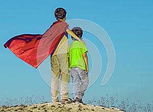 Two boys playing superheroes on the sky background, Superhero protects younger friend
