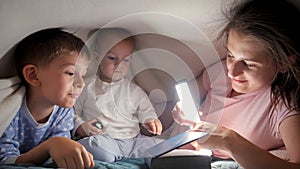 Two boys reading book with mother while lying under blanket at night. Family having time together, parenting, happy childhood and