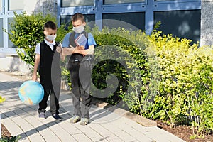 Two boys, schoolchildren in black school uniforms with backpacks in a medical mask go to school independently, hold hands,