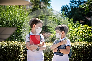 Two boys, schoolchildren in black school uniforms with backpacks in a medical mask go to school independently, hold hands,