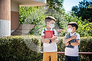 Two boys, schoolchildren in black school uniforms with backpacks in a medical mask go to school independently, hold hands,