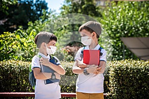 Two boys, schoolchildren in black school uniforms with backpacks in a medical mask go to school independently, hold hands,