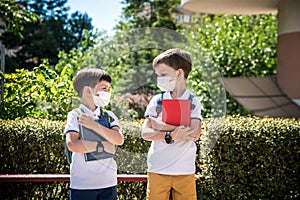 Two boys, schoolchildren in black school uniforms with backpacks in a medical mask go to school independently, hold hands,