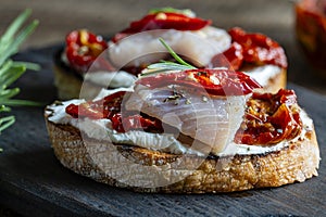 Two bread slices with ricotta cheese, pieces of herring and sun dried red tomatoes on wooden board background. Delicious breakfast