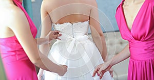 Two bride-maids in fuchsia dresses bustling the wedding dress. Bride-maids hands ribbon making on the back of bride white dress