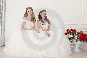 Two brides in wedding dresses sit on a white couch