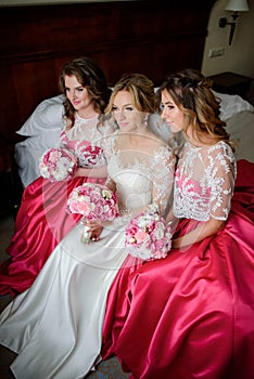 Two bridesmaids in pink dresses and stunning blonde bride sit on the bed
