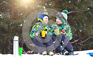 Two brothers drink hot tea in a snowy forest after sledging. Winter fun for Christmas vacation.