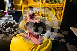 Two brothers playing video game console, sitting on yellow pouf in kids play center