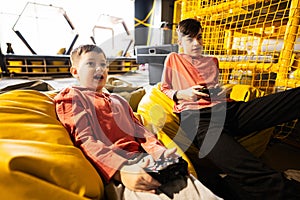 Two brothers playing video game console, sitting on yellow pouf in kids play center