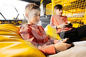 Two brothers playing video game console, sitting on yellow pouf in kids play center