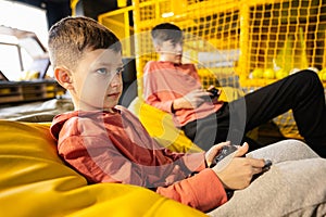 Two brothers playing video game console, sitting on yellow pouf in kids play center