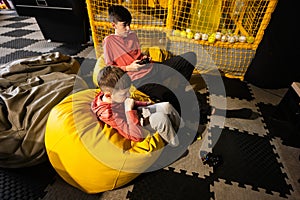 Two brothers playing video game console, sitting on yellow pouf in kids play center