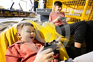 Two brothers playing video game console, sitting on yellow pouf in kids play center