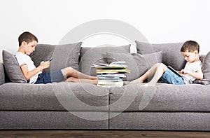 Two brothers sits in the opposite side of the sofa and looks on tablet computers.