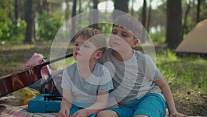 Two brothers sitting on picnic blanket and watching the fire with smoke during summer family camping vacation with tent