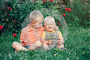 Two brothers sitting together on grass in park outside sharing apple. Older brother hug calm down soothe pacify crying younger