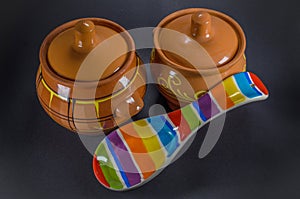 Two brown clay pots with colored ceramic spoon on black background