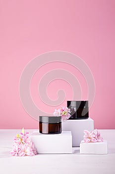 Two brown cosmetics containers for cream on white podium with fresh spring flowers on soft light pastel pink wall, vertical.