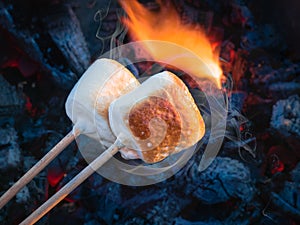 Two brown sweet marshmallows roasting over fire flames. Marshmallow on skewers roasted on charcoals