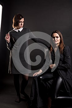 Two brunette girls in dark clothes in a photo studio on a dark background. One is sitting on a chair looking at the