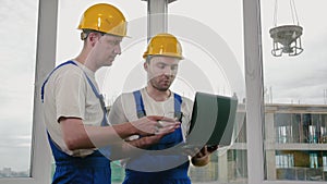 Two builders wearing protective helmets working on a laptop.