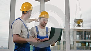 Two builders wearing protective helmets working on a laptop.