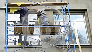 Two building contractors on scaffoldings are plastering, applying stucco finish, rendering, coating the facade wall of a house