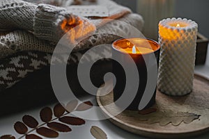 Two burning candles and stack of warm cozy sweaters on bedtable. Cozy autumn evening at home