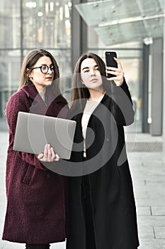 Two business girls take a selfie on the phone, work on a laptop on the background of the office, on the street