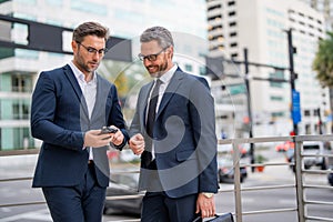 Two business men using mobile phone app texting outside of office in urban city. Businessmen holding smartphone for