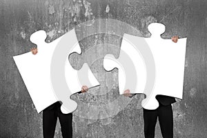 Two business people assembling white jigsaw puzzles with concrete wall photo