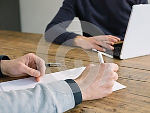 Two business people have meeting round a wooden table at an office. One writing one typing on laptop