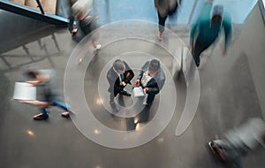 Two business people standing in the lobby of an office looking at a tablet while people are walking past in a blur