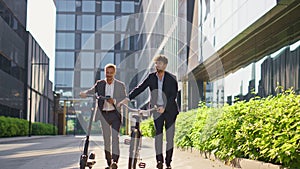 Two businessmen going city downtown. Smiling colleagues talking holding scooter