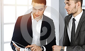 Two businessmen and partners working with tablet computer at the desk in sunny office. Multi ethnic teamwork