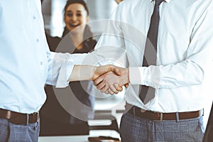 Two businessmen are shaking hands in office, close-up. Happy and excited business woman stands with raising hands at the