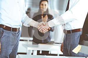 Two businessmen are shaking hands in office, close-up. Happy and excited business woman stands with raising hands at the