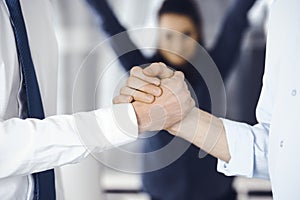 Two businessmen are shaking hands in office, close-up. Happy and excited business woman stands with raising hands at the