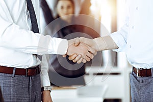 Two businessmen shaking hands in sunny office, close-up. Happy and excited business woman stands with raising hands at