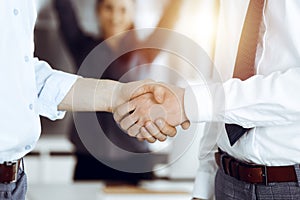 Two businessmen shaking hands in sunny office, close-up. Happy and excited business woman stands with raising hands at