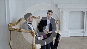 Two businessmen in suits with bow ties are communicating in a beautiful white room with elegant gold sofa.