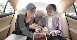 Two businesswoman sitting together in the backseat of car with digital tablet and talking.