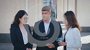 Two businesswomen and businessman, in jackets stand outdoor near office building and discuss the project, review the