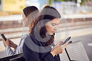 Two Businesswomen Commuting To Work Waiting For Train On Station Platform Looking At Mobile Phones