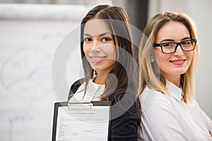 Two businesswomen coworkers standing in an office and smiling positively at the camera while holding folder of paperwork